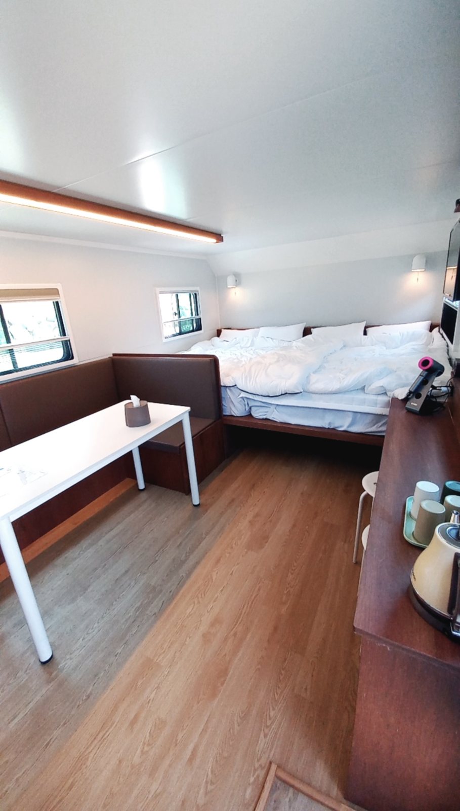 송1호 카라반 안 — 안쪽 침대와 소파형 의자, 흰 테이블, 나무 바닥